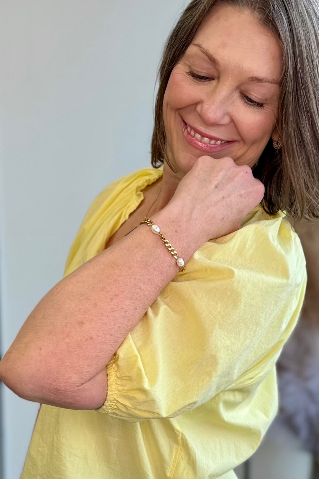 Woman wearing a yellow blouse smiling and looking down at her gold and pearl chain bracelet.