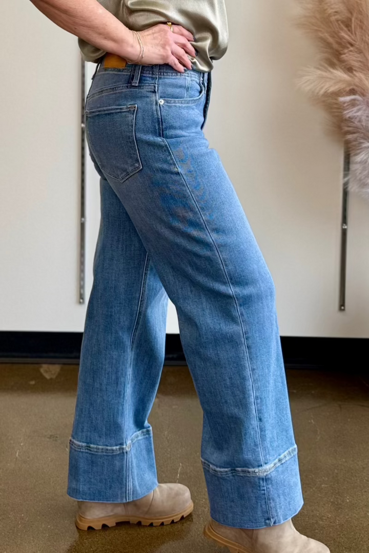 Side close up view of a medium wash jean with a wide cuff worn by a woman standing with her hands on her hips.