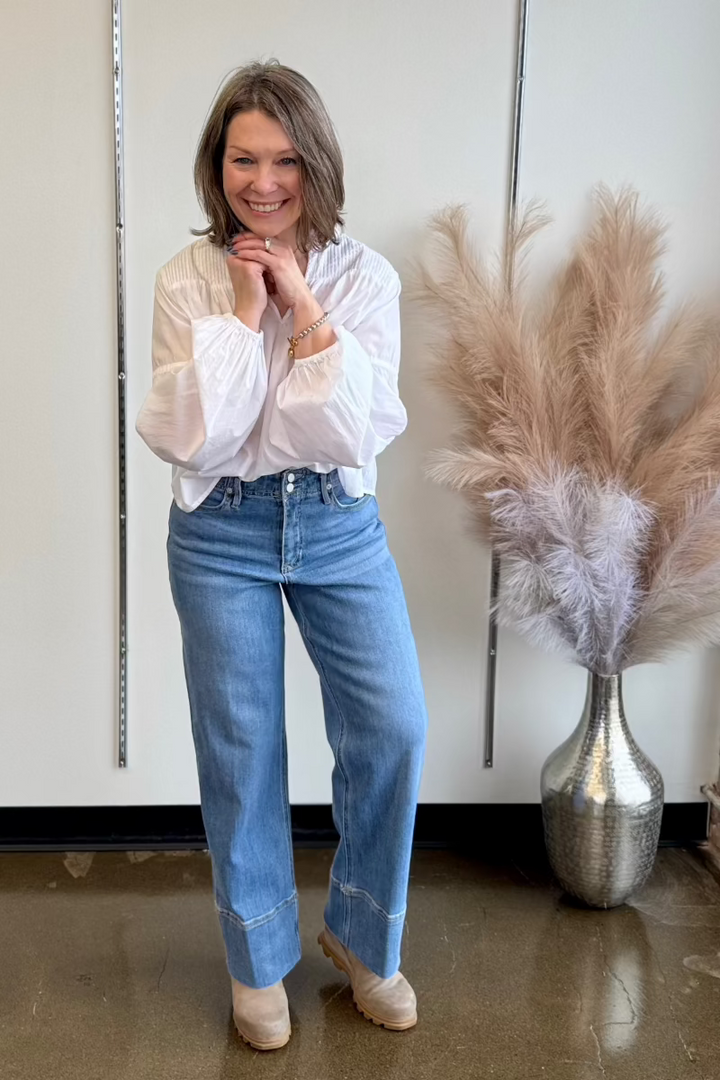 Photo of smiling woman holding her hands under her chin, wearing a white flowy blouse and medium wash jeans.