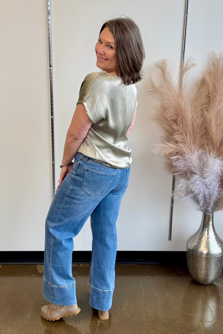 Woman smiling and looking over her shoulder wearing a silky short sleeve neutral blouse and jeans.