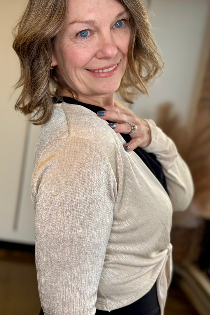 Close up side view of a woman wearing a front tie light gold top with her hand on her chest.