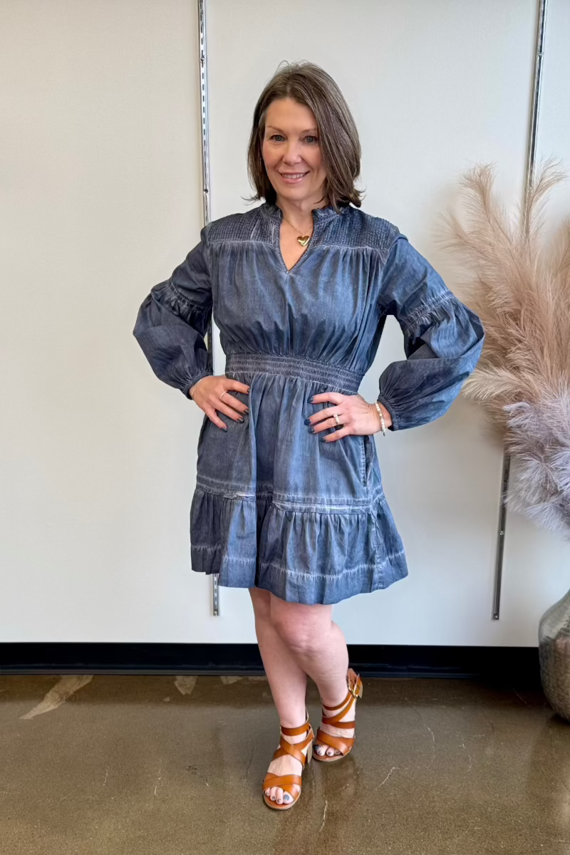 Woman wearing a long sleeve blue dress and brown sandals standing with her hands on her hips.