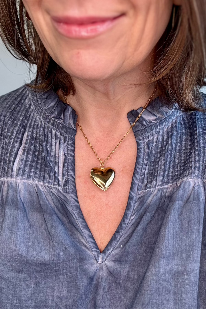 Close up of a smooth gold heart necklace worn by a woman wearing a blue v-neck.